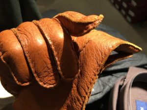A brown leather glove, worn by a person, is prominently displayed, highlighting its texture and stitching under bright lighting in an indoor setting.